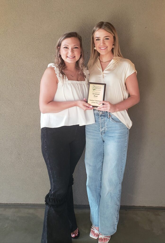 Two young ladies standing with one holding a volunteer award plaque.
