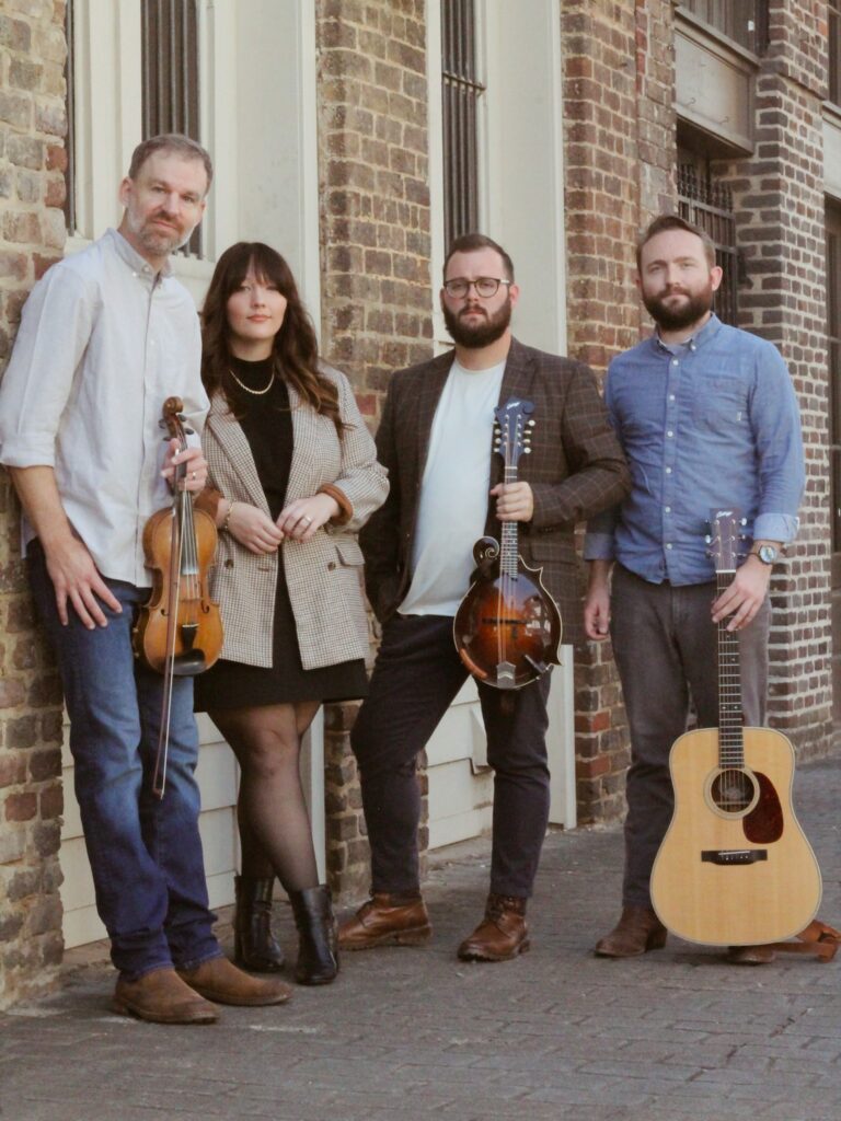 Four adults leaning against a wall. The woman has a violin. One man has a guitar. Cawling band.

